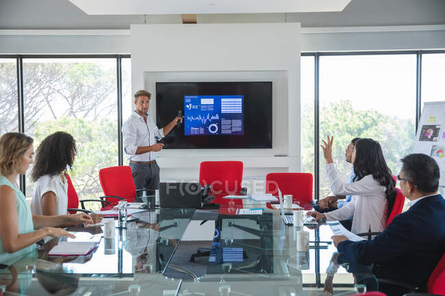Kaukasische Geschäftsfrau, die neben einem Bildschirm steht und männlichen und weiblichen Kollegen im Besprechungsraum eine Präsentation gibt, wobei eine ihre Hand hebt. Kreative Geschäftsprofis, die in einem modernen Büro arbeiten. — Stockfoto