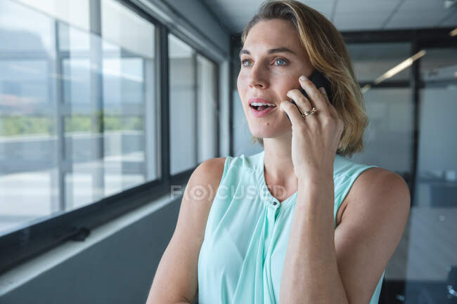 Умная, непринужденно одетая белая деловая женщина, творческая женщина с светлыми волосами, выглядывающая в окно, разговаривающая по смартфону. Креативный бизнес-профессионал, работающий в современном офисе. — стоковое фото
