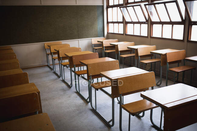 Vue latérale d'une salle de classe vide, avec des bureaux en formation et un tableau noir derrière eux, avec des fenêtres ouvertes par temps ensoleillé — Photo de stock