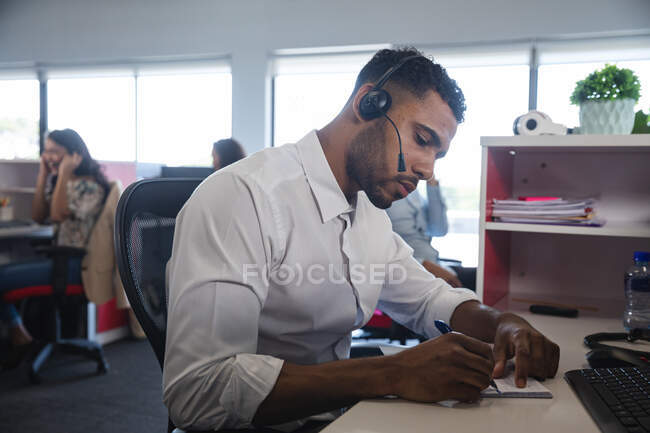 Smart casually dressed mixed race businessman sitting at desk wearing phone headset and writing notes, with colleagues in the background. Creative business professionals working in busy modern office. — Stock Photo