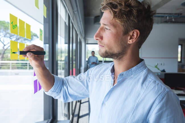 Smart casually dressed Caucasian businessman writing memos on sticky notes stuck to a window. Creative business professional working in a busy modern office. — Stock Photo