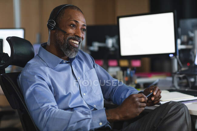 African American businessman working late in the evening in a modern office, sitting at a desk, wearing a phone headset and smiling. — Stock Photo