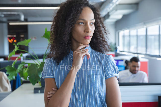 Smartly dressed mixed race female business creative with long hair looking away with hand on chin, a colleague working in the background. Creative business professionals working in a modern office. — Stock Photo