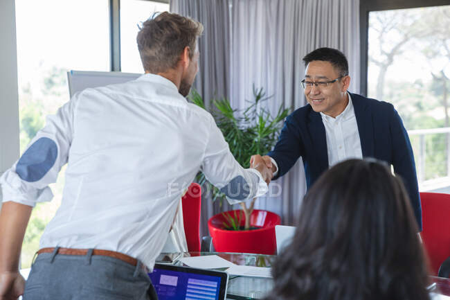 Homme d'affaires asiatique et caucasien serrant la main à travers la table lors d'une réunion d'affaires, avec des collègues assis à côté d'eux. Professionnels créatifs d'affaires travaillant dans un bureau moderne occupé. — Photo de stock