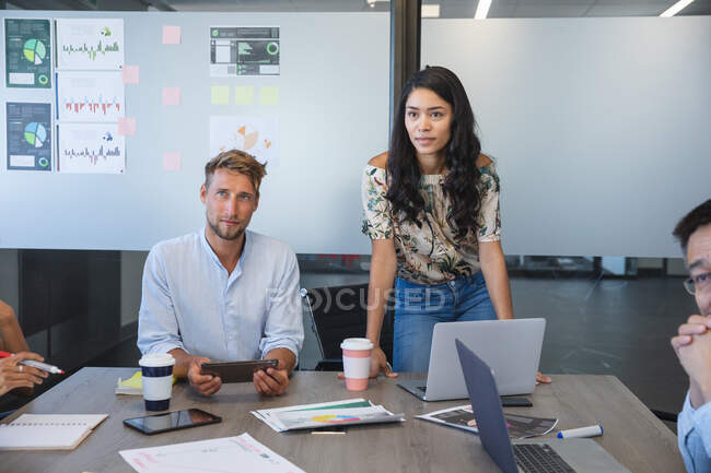 Asiatische Geschäftsfrau, die während einer Teamsitzung vor Kollegen steht, ein kaukasischer männlicher Kollege, der neben ihr sitzt und zuhört. Kreative Geschäftsprofis, die in einem modernen Büro arbeiten. — Stockfoto