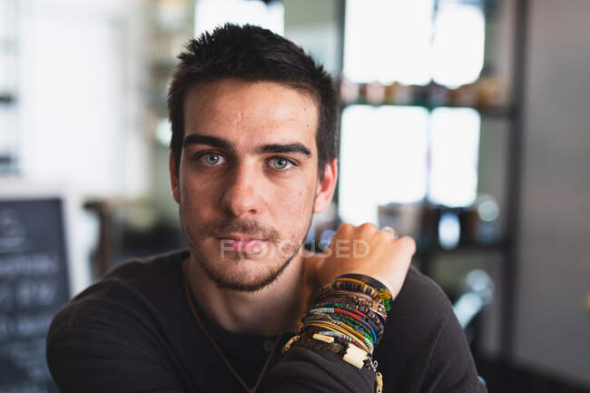 Portrait of a Caucasian man wearing casual clothes and jewelry, sitting by a table in a coffee shop, looking straight into a camera — Stock Photo