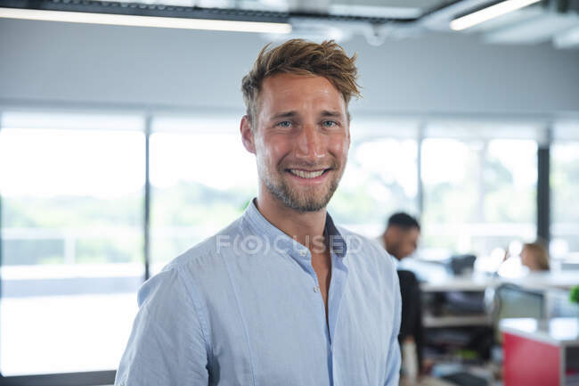 Porträt eines selbstbewussten kaukasischen Geschäftsmannes mit kurzen Haaren und Bart, der lächelt, während im Hintergrund Kollegen arbeiten. Kreativer Geschäftsprofi, der in einem geschäftigen modernen Büro arbeitet. — Stockfoto