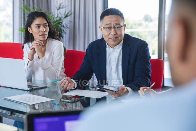 Hombre de negocios asiática mayor sosteniendo tableta y hablando durante una reunión de negocios, una colega asiática sentada a su lado escuchando. Profesionales creativos de negocios que trabajan en una oficina moderna ocupada. - foto de stock