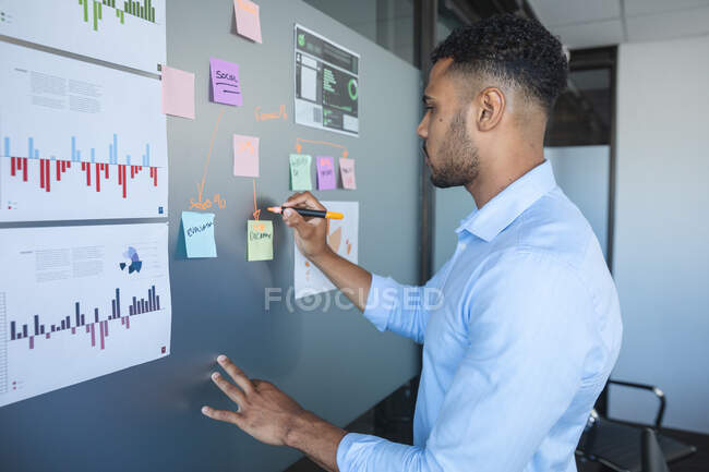 Homem de negócios misto casualmente vestido inteligente escrevendo notas em um quadro magnético com gráficos e informações sobre ele. Profissional de negócios criativos trabalhando em um escritório moderno ocupado. — Fotografia de Stock
