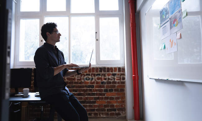 Uomo caucasico che lavora in un ufficio informale, usando il suo portatile e guardando a bordo con piani. Distanze sociali sul luogo di lavoro durante la pandemia di Coronavirus Covid 19. — Foto stock