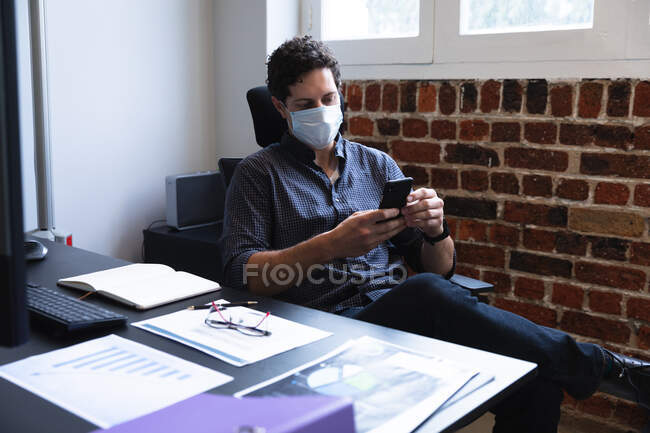 Uomo caucasico che lavora in un ufficio informale, usando il suo smartphone e indossando maschera facciale. Distanze sociali sul luogo di lavoro durante la pandemia di Coronavirus Covid 19. — Foto stock
