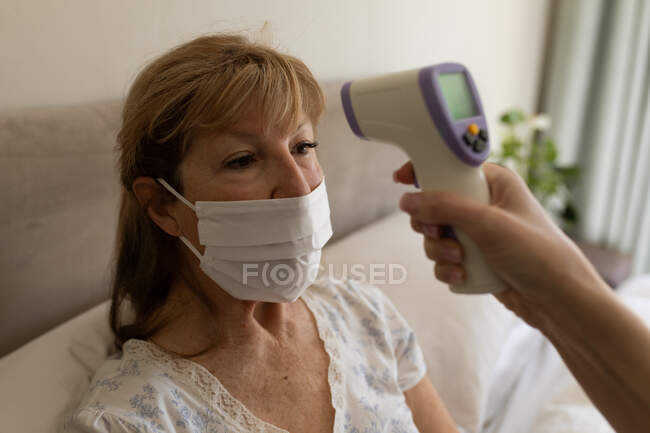 Mujer mayor caucásica en casa visitada por enfermera caucásica, comprobando la temperatura. Atención médica en el hogar durante la cuarentena del Coronavirus de Covid 19. - foto de stock