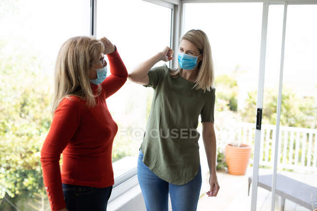 Donna caucasica anziana e sua figlia adulta a casa, indossano maschere facciali e si salutano toccandosi i gomiti. Distanze sociali, salute e igiene durante la pandemia di Covid 19 Coronavirus. — Foto stock