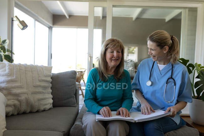 Старшая белая женщина дома, навещаемая белой медсестрой, читает книгу руками. Медицинская помощь на дому во время карантина Ковид 19 Коронавирус. — стоковое фото