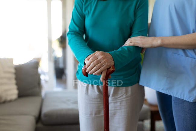 Femme à la maison visitée par une infirmière, debout avec une canne. Soins médicaux à domicile pendant la quarantaine du coronavirus Covid 19. — Photo de stock