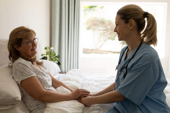Donna caucasica anziana a casa visitata da infermiera caucasica, seduta sul letto, che si tiene per mano. Assistenza medica a domicilio durante la quarantena di Covid 19 Coronavirus. — Foto stock