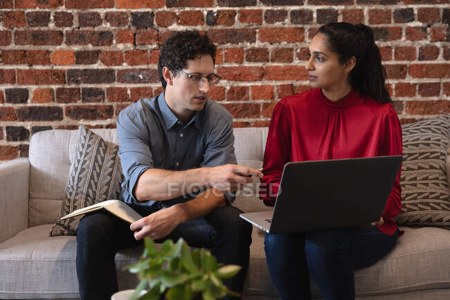 Femme métisse et homme caucasien travaillant dans un bureau décontracté, assis sur un canapé, utilisant un ordinateur portable et parlant. Professionnels créatifs d'affaires travaillant dans un bureau moderne occupé. — Photo de stock