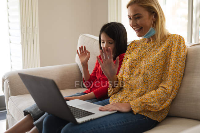 Femme caucasienne et sa fille passent du temps à la maison ensemble, portant des masques, utilisant un ordinateur portable, faire un appel vidéo. Distance sociale pendant le confinement en quarantaine du coronavirus Covid 19. — Photo de stock