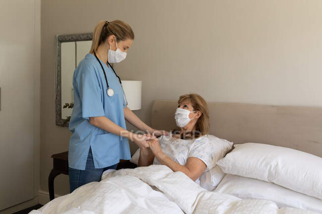 Mulher caucasiana sênior em casa visitada por enfermeira caucasiana, de pé e de mãos dadas. Cuidados médicos em casa durante a quarentena do Coronavirus Covid 19. — Fotografia de Stock