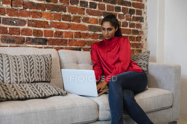 Gemischte Rasse Frau arbeitet in einem lässigen Büro, sitzt auf einem Sofa und benutzt einen Laptop-Computer. Soziale Distanzierung am Arbeitsplatz während der Coronavirus Covid 19 Pandemie. — Stockfoto