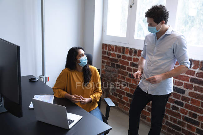 Mulher de raça mista e homem caucasiano trabalhando em um escritório casual, usando máscaras faciais e conversando. Distanciamento social no local de trabalho durante a pandemia do Coronavirus Covid 19. — Fotografia de Stock