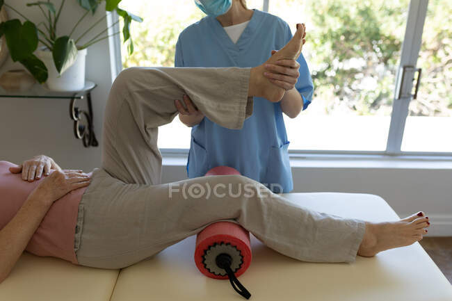 Donna caucasica anziana a casa visitata da infermiera caucasica, allungando la gamba, indossando una maschera facciale. Auto medica — Foto stock