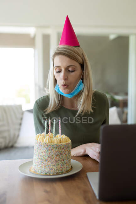 Kaukasische Frau verbringt Zeit zu Hause, sitzt im Wohnzimmer mit Geburtstagstorte und Laptop und bläst Kerzen. Soziale Distanzierung während Covid 19 Coronavirus Quarantäne. — Stockfoto