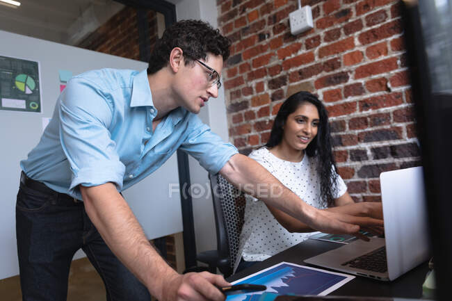 Frau mit gemischter Rasse und Mann aus dem Kaukasus arbeiten in einem lockeren Büro, benutzen einen Laptop und diskutieren über ihre Arbeit. Kreative Geschäftsprofis, die in einem modernen Büro arbeiten. — Stockfoto