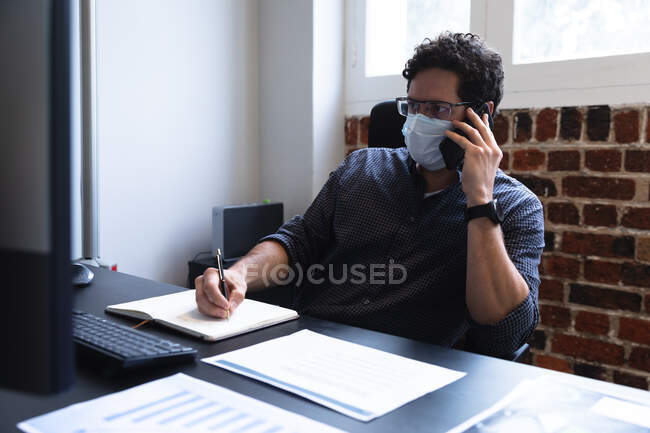 Homme caucasien travaillant dans un bureau décontracté, parlant sur smartphone, prenant des notes et portant un masque facial. Distance sociale sur le lieu de travail pendant la pandémie de coronavirus Covid 19. — Photo de stock