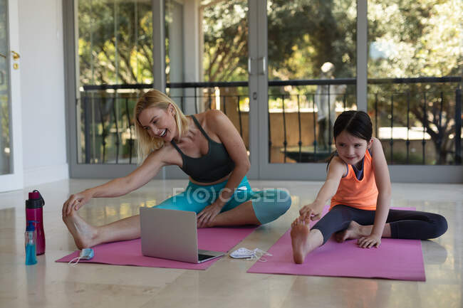 Femme caucasienne et sa fille passent du temps à la maison ensemble, faisant du yoga, s'étirant, utilisant un ordinateur portable. Distance sociale pendant le confinement en quarantaine du coronavirus Covid 19. — Photo de stock