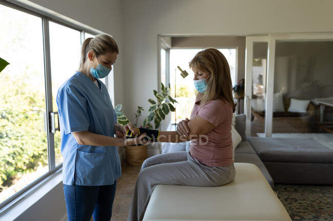 Mujer mayor caucásica en casa visitada por una enfermera caucásica, estirando su muñeca, usando máscaras faciales. Atención médica en el hogar durante la cuarentena del Coronavirus de Covid 19. - foto de stock