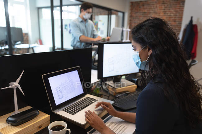 Homme caucasien et femme métisse travaillant dans un bureau décontracté, portant des masques faciaux, utilisant des ordinateurs portables. Distance sociale sur le lieu de travail pendant la pandémie de coronavirus Covid 19. — Photo de stock