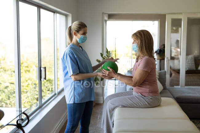 Senior Caucasian woman at home visited by Caucasian female nurse, doing exercises with a ball, wearing face masks. Medical car — Stock Photo