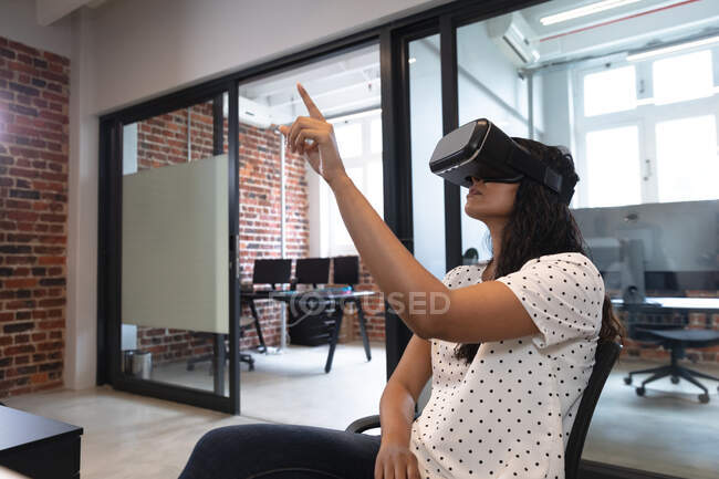 Femme de race mixte travaillant dans un bureau décontracté, portant un casque vr, un écran virtuel tactile. Distance sociale sur le lieu de travail pendant la pandémie de coronavirus Covid 19. — Photo de stock