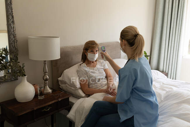 Femme caucasienne senior à la maison visité par une infirmière caucasienne, vérifier la température. Soins médicaux à domicile pendant la quarantaine du coronavirus Covid 19. — Photo de stock