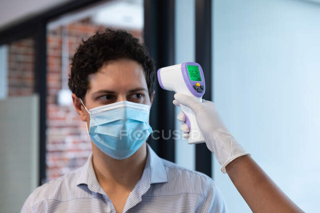 Mixed race woman and Caucasian man working in a casual office, wearing face mask, a woman taking his temperature. Social distancing in the workplace during Coronavirus Covid 19 pandemic. — Stock Photo