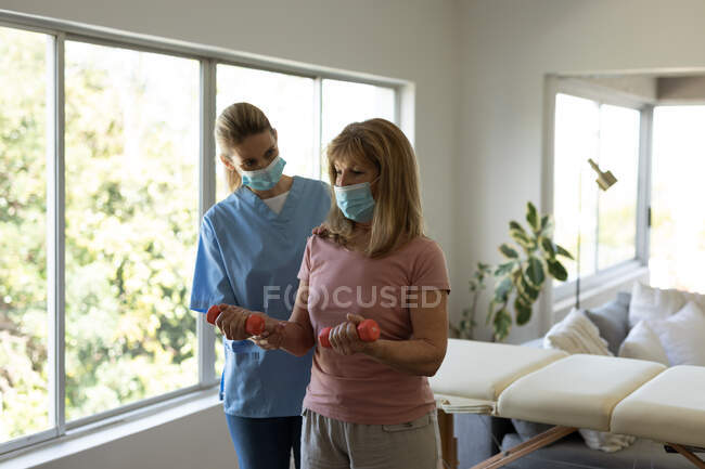 Senior Caucasian woman at home visited by Caucasian female nurse, stretching her arm, wearing face masks. Medical care at home during Covid 19 Coronavirus quarantine. — Stock Photo