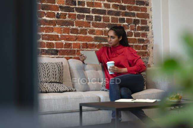 Donna di razza mista che lavora in un ufficio informale, seduta su un divano, usando un tablet. Distanze sociali sul luogo di lavoro durante la pandemia di Coronavirus Covid 19. — Foto stock