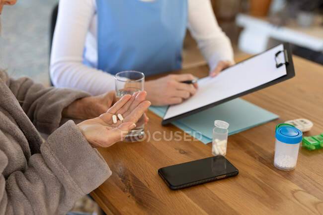 Donna a casa visitata da infermiera donna, seduta al tavolo con in mano una cartellina, donna che prende pillole. Assistenza medica a domicilio durante la quarantena di Covid 19 Coronavirus. — Foto stock