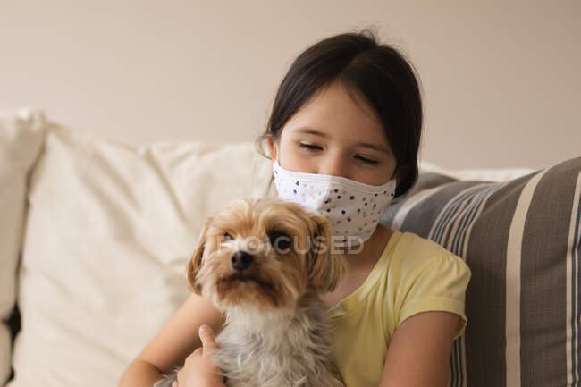 Caucasian girl spending time at home, wearing face mask, embracing her dog. Social distancing during Covid 19 Coronavirus quarantine lockdown. — Stock Photo