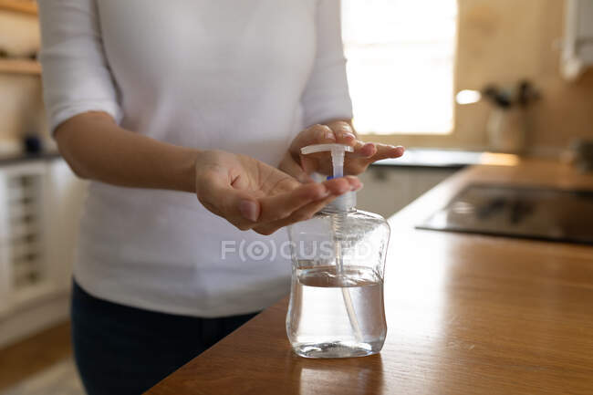 Mujer pasando tiempo en casa, de pie en la cocina desinfectando sus manos. Distanciamiento social e higiene durante la cuarentena del Coronavirus Covid 19. - foto de stock