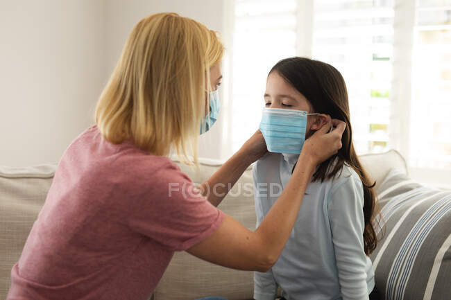 Donna caucasica e sua figlia passano del tempo a casa insieme, madre che aiuta la figlia a mettersi la maschera facciale. Distanza sociale durante il blocco di quarantena Covid 19 Coronavirus. — Foto stock