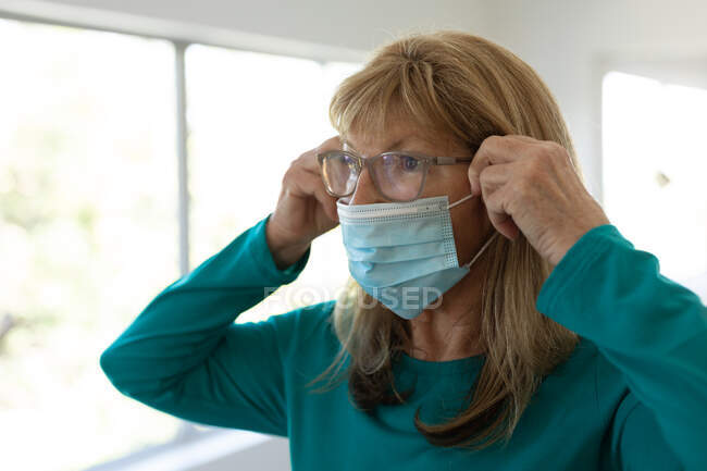Старшая белая женщина проводит время дома, стоит в гостиной и надевает маску для лица. Социальное дистанцирование во время изоляции коронавируса Covid 19. — стоковое фото