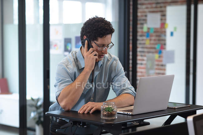 Белый мужчина работает в обычном офисе, разговаривает на смартфоне и пользуется ноутбуком. Социальное дистанцирование на рабочем месте во время пандемии Coronavirus Covid 19. — стоковое фото
