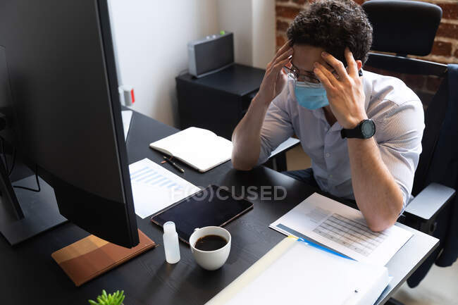 Kaukasischer Mann, der in einem lässigen Büro arbeitet, seine Stirn berührt und eine Gesichtsmaske trägt. Soziale Distanzierung am Arbeitsplatz während der Coronavirus Covid 19 Pandemie. — Stockfoto