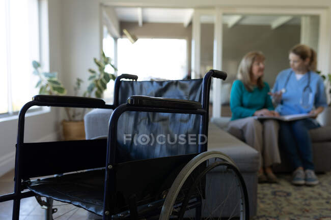 Mulher branca sênior em casa visitada por enfermeira caucasiana, sentada no sofá, conversando com uma cadeira de rodas em primeiro plano. Cuidados médicos em casa durante a quarentena do Coronavirus Covid 19. — Fotografia de Stock
