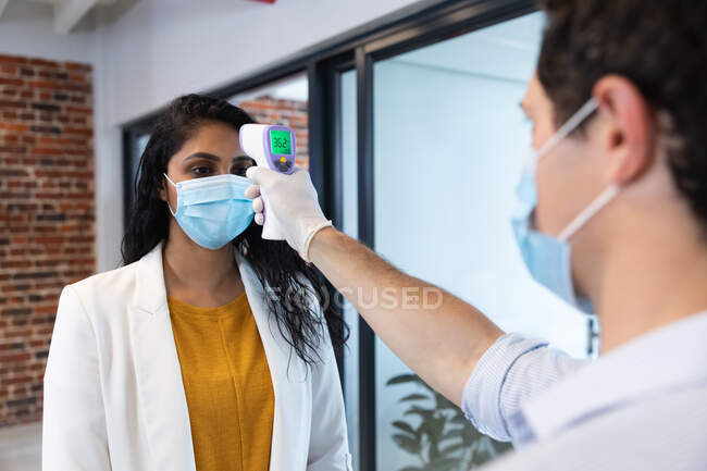Смешанная расовая женщина и белый мужчина, работающий в обычном офисе, в масках, мужчина, измеряющий ее температуру. Социальное дистанцирование на рабочем месте во время пандемии Coronavirus Covid 19. — стоковое фото