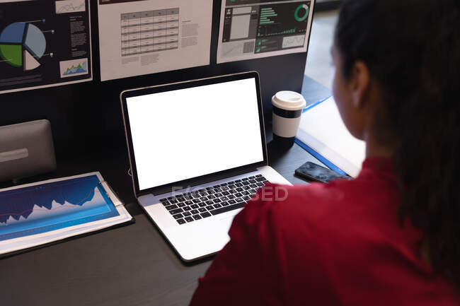 Femme de race mixte travaillant dans un bureau décontracté, assise au bureau, à l'aide d'un ordinateur portable. Distance sociale sur le lieu de travail pendant la pandémie de coronavirus Covid 19. — Photo de stock
