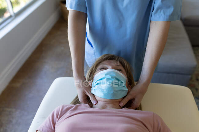 Mulher branca sênior em casa visitada por enfermeira caucasiana, massageando o pescoço, usando máscara facial. Carro médico — Fotografia de Stock
