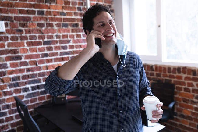 Белый мужчина работает в офисе, разговаривает по смартфону, держит кофе на вынос и носит маску для лица. Социальное дистанцирование на рабочем месте во время пандемии Coronavirus Covid 19. — стоковое фото